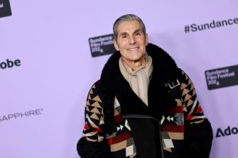 Smiling older man with short gray hair wearing a patterned coat and beige scarf posing in front of a light purple Sundance Film Festival backdrop.