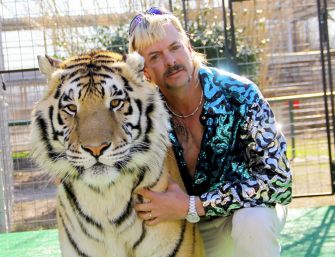 Man with a mustache posing beside a tiger in a fenced enclosure.
