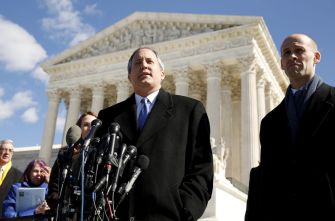 A man in a suit speaks at a press conference in front of the Supreme Court building, flanked by supporters and microphones.