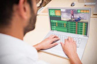 Man viewed from behind using a laptop displaying a sports betting interface with a basketball game and odds.