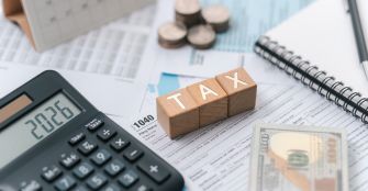 Close-up of a calculator, stacks of coins, dollar bills, and wooden blocks spelling TAX on tax documents.