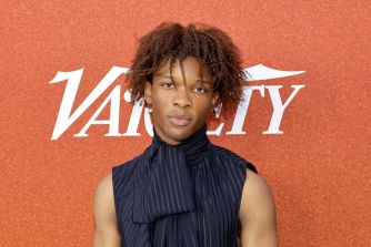 Close-up portrait of a young man with short dreadlocks in a dark, vertically striped shirt, standing in front of an orange background with white lettering.