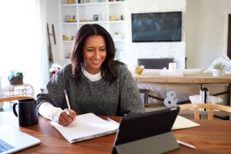 A woman sitting at a table, working on a notepad while using a tablet and laptop in a well-lit home environment.