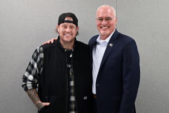 Two men standing side by side and smiling for the camera; one wears a black cap and plaid shirt, the other wears a dark suit.