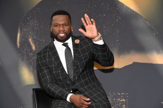 Man in a black checkered suit waving at a formal event with a gold-toned backdrop