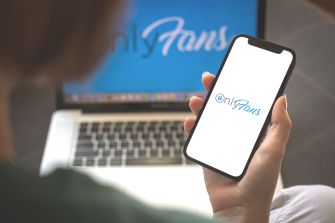 A person holding a smartphone displaying the OnlyFans logo, with a laptop in the background.