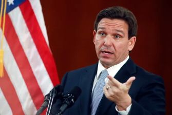 Man in a suit gestures while speaking at a podium, with an American flag in the background.