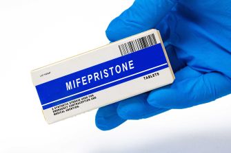 A blue-gloved hand holding a box labeled Mifepristone (the abortion pill).