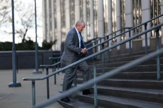 A man in a gray suit walks down outdoor steps beside a railing outside a courthouse.