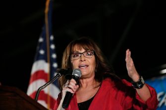 Woman in a red blazer and glasses speaks at a podium with a microphone, American flag visible in the background.