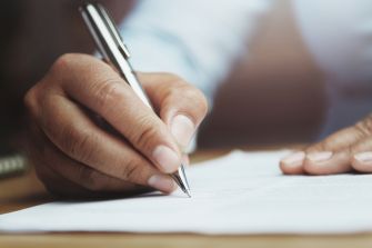 A hand holding a pen is seen writing on a sheet of paper, symbolizing the signing of a contract.