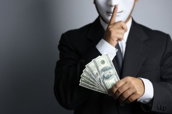 A person in a suit with a mask holds a finger to their lips while displaying cash, symbolizing secrecy in campaign finance.