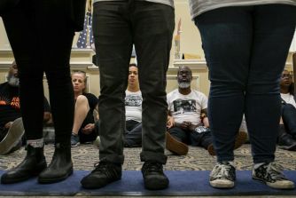A group of individuals sitting in protest, focusing on their legs and shoes, with a backdrop of flags and governmental decor.