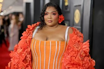 Lizzo posing on the red carpet at an event, wearing a vibrant orange outfit with floral accents.