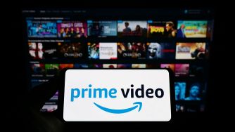 A smartphone displaying the Prime Video logo in front of a blurred TV screen with various movie and show thumbnails.