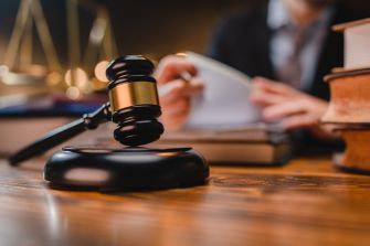 Close-up of a judge's gavel resting on a wooden desk with legal books nearby and a blurred figure in a suit in the background.