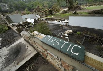 Debris and a broken wall with a green sign reading “MYSTIC” amid flood-damaged structures at Camp Mystic.