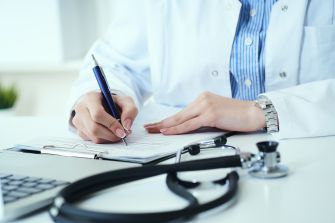 A doctor writing notes, with a stethoscope and computer in the foreground.