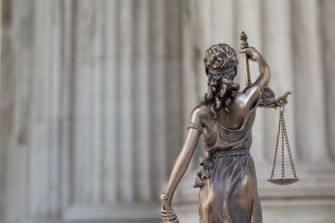 A bronze statue of Lady Justice holding scales, viewed from behind, set against a classical architectural backdrop.