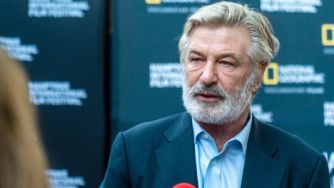 Alec Baldwin speaking at an event, with a backdrop featuring the National Geographic logo.