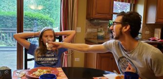 A candid moment at a dining table with two people sharing a lighthearted interaction. One person is playfully offering a drink to another, while the other looks amused and relaxed. The setting features a cozy kitchen with natural light and a view of the outdoors.