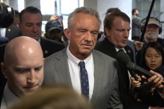 Man in a gray suit being interviewed by reporters, surrounded by microphones and photographers.