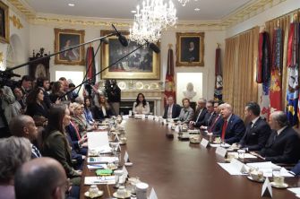 A meeting taking place at a conference table, with several individuals seated on either side, and cameras capturing the event, set in an elaborately decorated room.