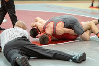 Two male wrestlers grappling on a wrestling mat as a striped-shirt referee watches nearby.