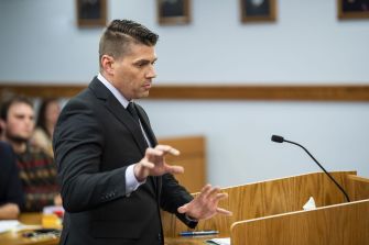 A man in a suit gestures while speaking at a courtroom podium, with a blurred audience visible in the background.