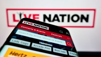 Close-up of a smartphone displaying the Live Nation app, with a blurred Live Nation sign in the background and “Concerts & Events” visible on the screen.