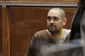 Man in jail uniform seated in a courtroom, with wood-paneled walls in the background.