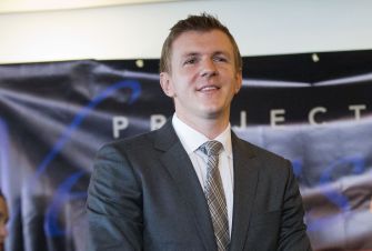 A man in a suit stands in front of a Project Veritas banner, appearing confident during a public event.