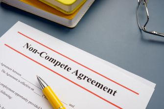 A close-up of a non-compete agreement document on a desk, accompanied by a yellow pen and reading glasses.