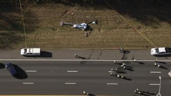Aerial view of a helicopter crash site alongside an interstate highway, with emergency responders at the scene.