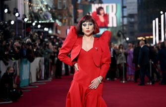 Lady Gaga posing on a red carpet in a bright red dress with dramatic shoulder details, photographers and fans visible in the background.