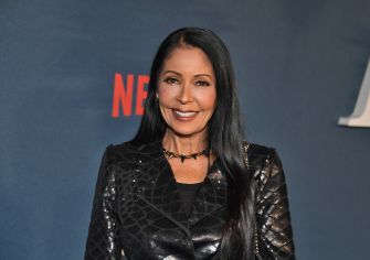 Portrait of a smiling woman with long black hair in a sequined black outfit, standing in front of a blue backdrop.