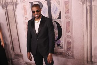Man in a black suit and sunglasses posing at an event with a patterned wall backdrop.