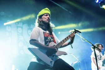 Guitarist with long hair wearing a bright lime beanie, playing a white V-shaped guitar on stage under blue and yellow lights.