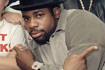 Close-up of a man wearing a black fedora and gray jacket, with his arms crossed and looking at the camera.