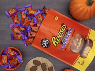 A bag of Reese's Pumpkin-shaped candies with a few scattered on a tabletop alongside pumpkins and other candy.