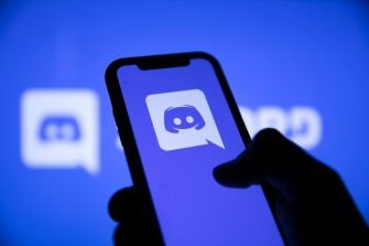 A hand holding a smartphone displaying the Discord logo against a blue background.