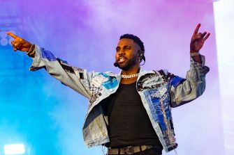 Male singer on stage with arms raised, wearing a patchwork denim jacket and black shirt, headset microphone, under colorful blue and purple lighting.