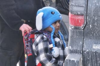 Young child wearing a blue knit hat with white ear flaps and a plaid jacket stands beside a snow-covered vehicle as an adult offers support.