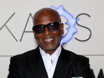 Portrait of a smiling, bald man in a black suit and white shirt wearing dark sunglasses, with a blue gear-like logo in the background.
