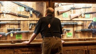 A man stands in a gun shop, looking at rifles displayed on wooden shelves.