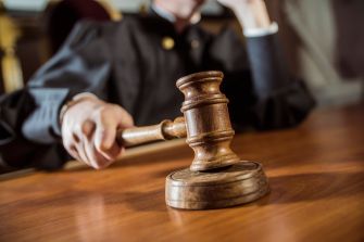 Gavel resting on a wooden judge's bench in a courtroom with a blurred robed figure in the background.