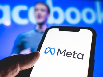 A person holding a phone displaying the Meta logo, with a blurred background of a speaker on stage.
