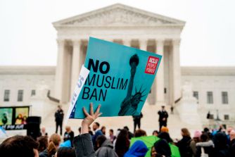 A protestor holding a sign that reads "No Muslim Ban" in front of the Supreme Court building, with a crowd gathered around.