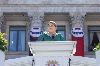 Woman at an outdoor podium giving a speech, with patriotic red, white, and blue bunting in the background.