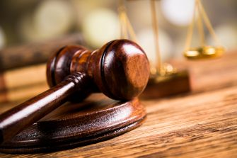 Image of a wooden gavel resting on a bench with a blurred background featuring justice scales.